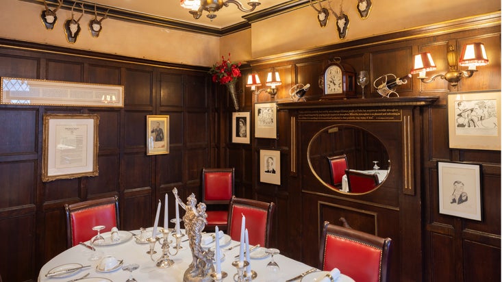 Intimate wood-panelled dining room featuring red leather chairs, a round mirror, framed illustrations and photographs, decorative wall lighting, and a formally set round table with silver candelabras.